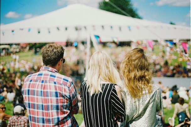 The backs of three people who look out over an outdoor event while the sun shines The backs of three people who look out over an outdoor event while the sun shines