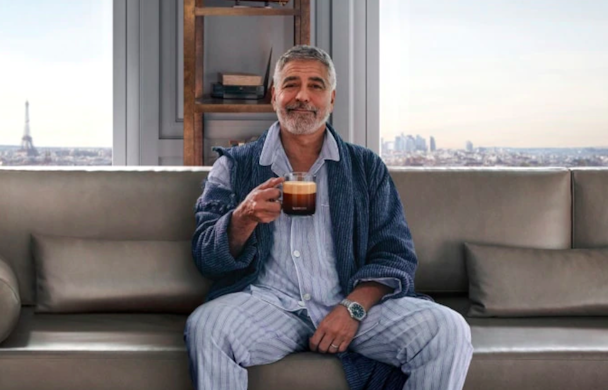 George Clooney sits on a sofa holding a cup of black coffee George Clooney sits on a sofa holding a cup of black coffee